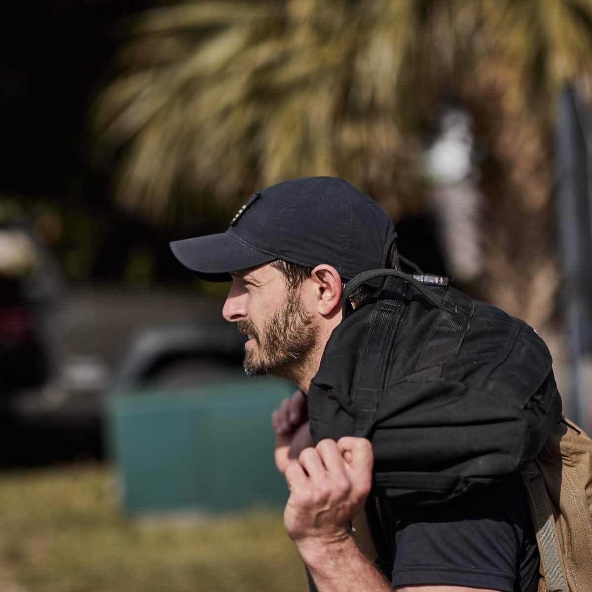 A man wearing a Performance TAC Hat - TOUGHDRY by GORUCK carries a black backpack over his shoulder. He is outdoors, with palm trees and a park-like setting in the background.