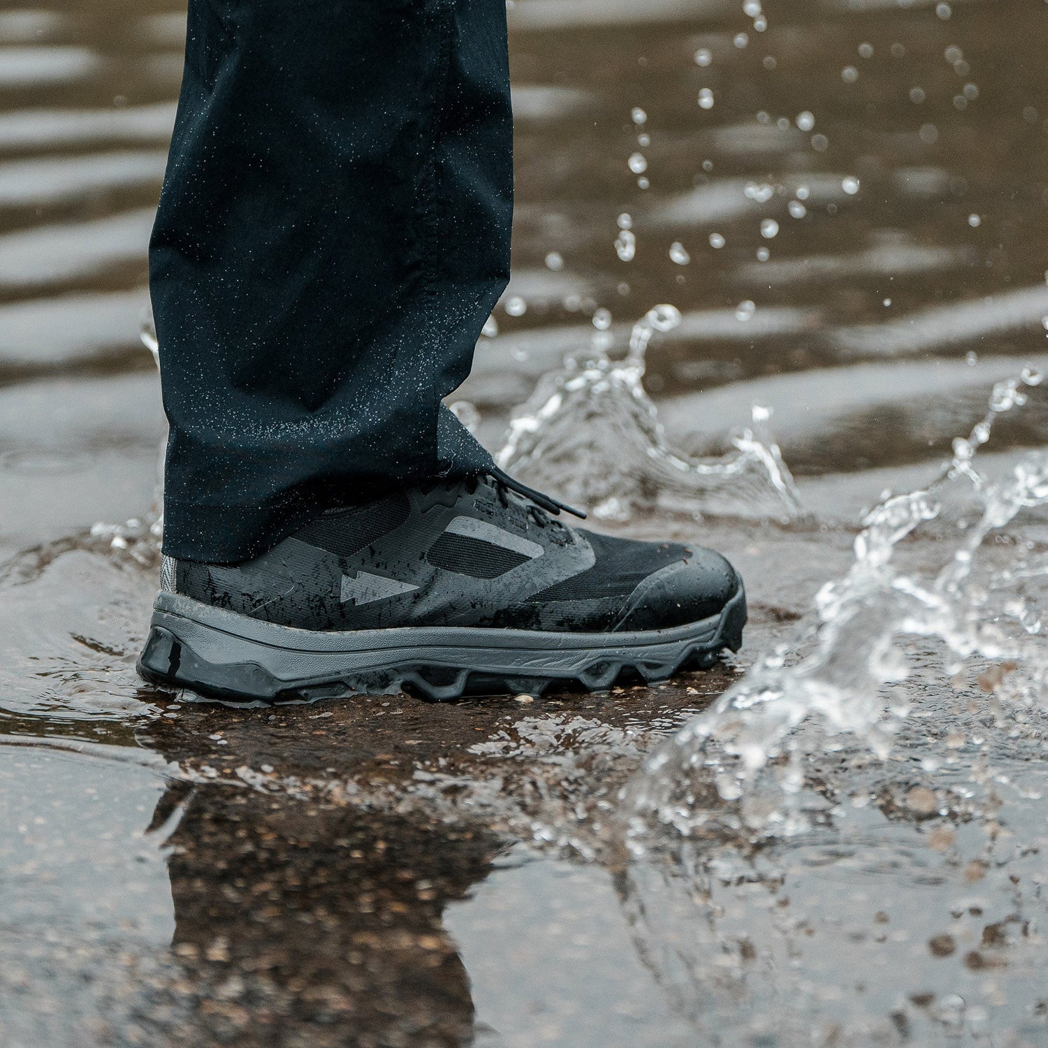 A person’s foot in a black waterproof shoe steps in a puddle, splashing water.