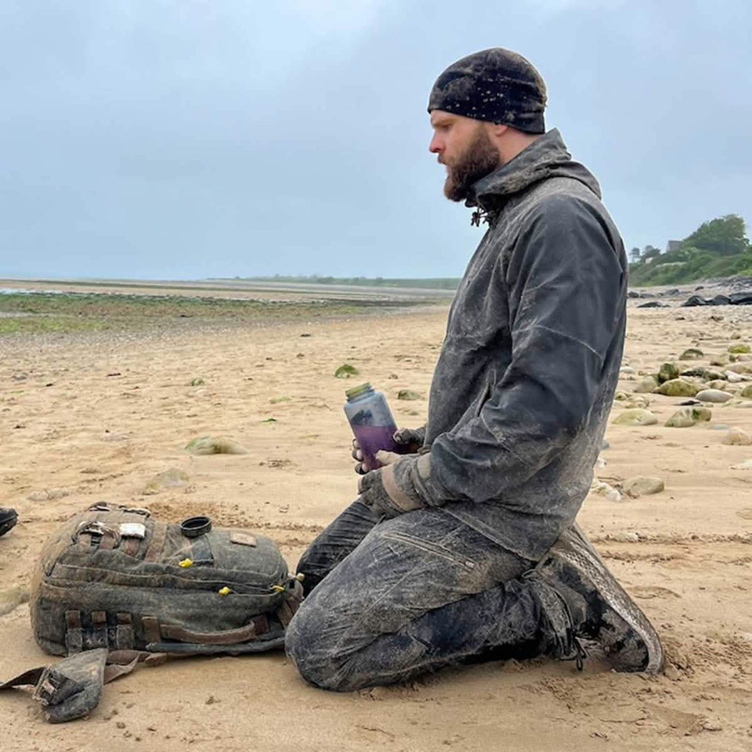 A person kneels on a sandy beach, wearing a hat and rugged clothing made from abrasion-resistant fabric. Holding a container, their Men’s Challenge Pants - Lightweight ToughDry® by GORUCK are ideal for the setting. A backpack rests beside them against the backdrop of a cloudy sky and expansive shoreline.