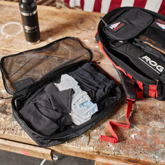 An open backpack on a wooden table features a mesh compartment containing folded clothing and a cloth, all neatly organized with GORUCK Packing Cubes. A water bottle rests nearby, while part of an American flag waves gently in the background. These packing cubes are your perfect travel companion.