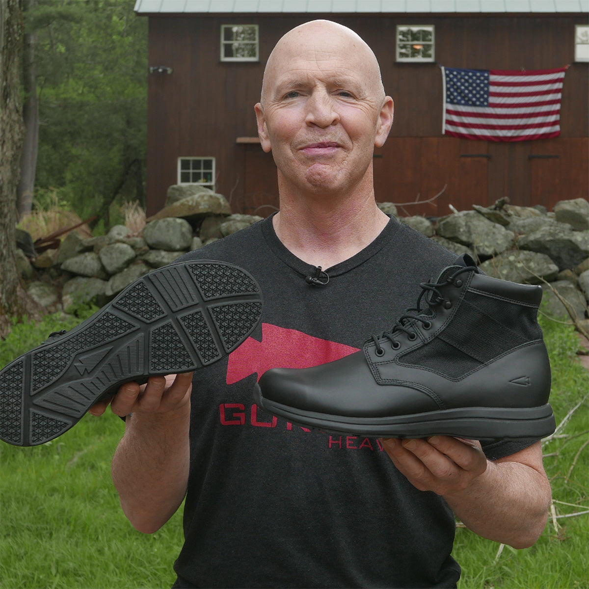 Bald man outdoors holding a black boot and its sole, with a barn and American flag in the background.