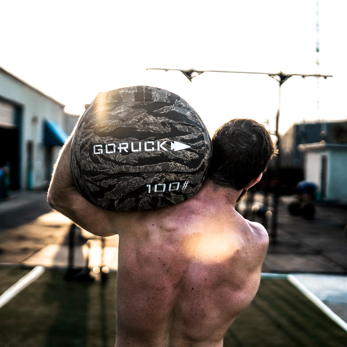 Shirtless person lifting a 100-pound GORUCK sandbag outdoors in a workout area at sunset.