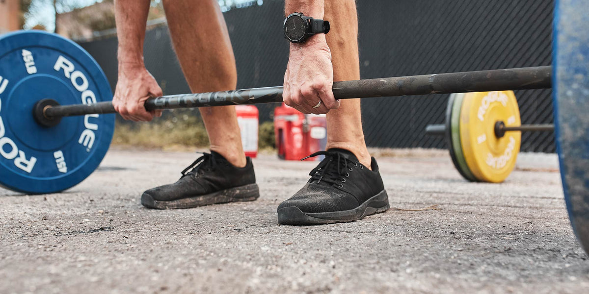 Person wearing black shoes gripping a barbell with blue and yellow weights, preparing to lift outdoors.