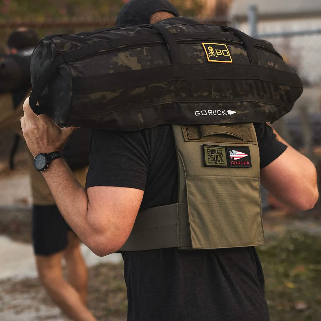 A person, wearing a GORUCK Training Weight Vest 2.0 with patches, carries a black sandbag over their shoulders and uses ruck plates for added intensity.