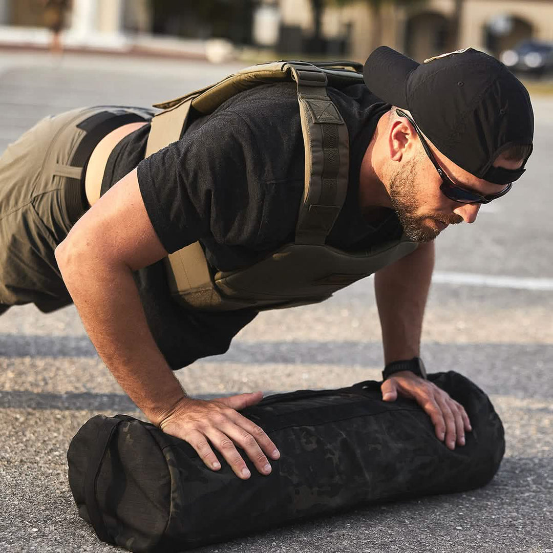A man in a GORUCK Training Weight Vest 2.0 and a tactical cap does push-ups with a weighted sandbag in a parking lot.