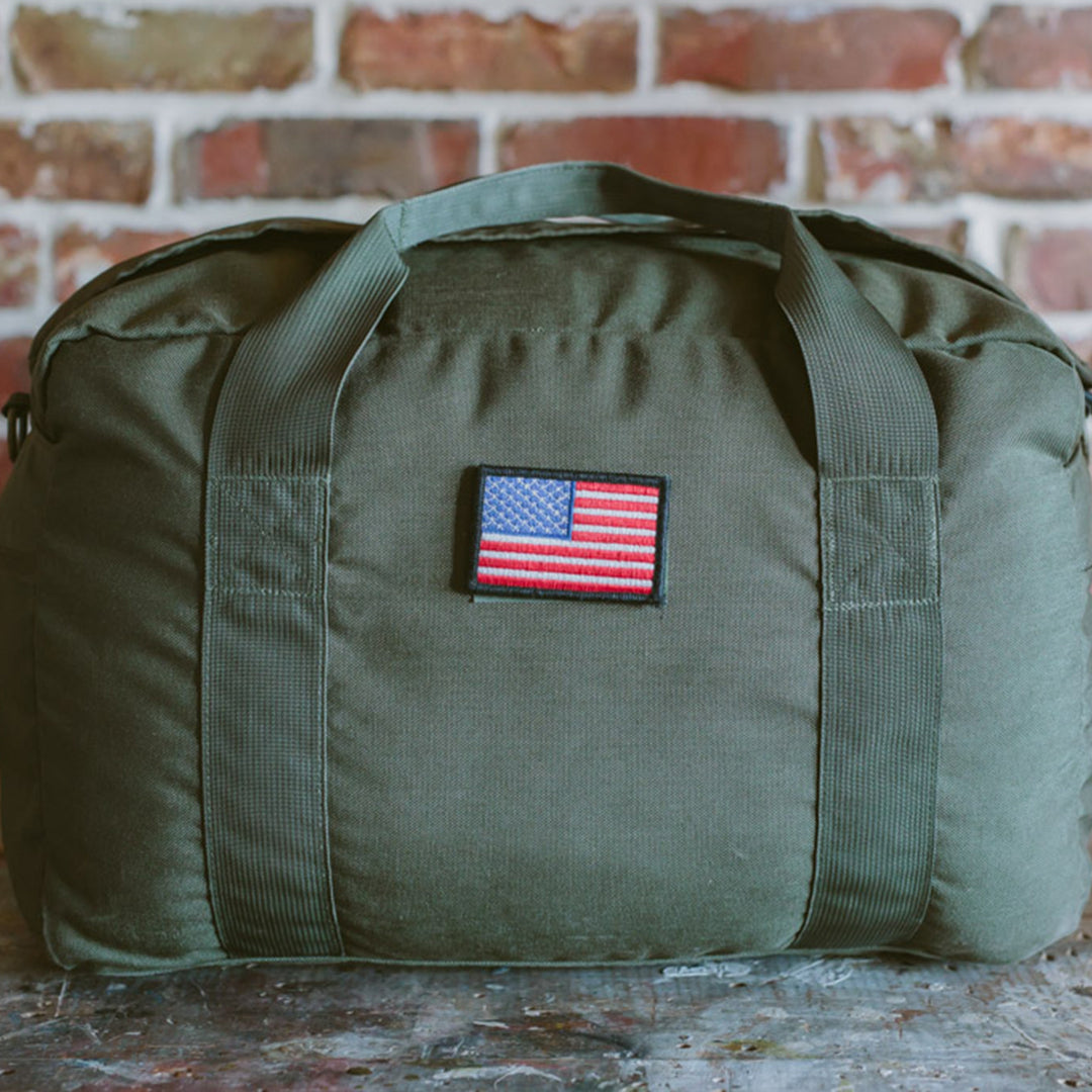 The GORUCK Kit Bag, in an olive green hue and featuring an American flag patch, evokes the design of a Special Forces Flight Bag and is displayed on a stone surface with a brick wall backdrop.