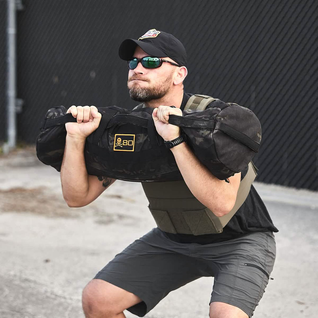 A man squats outdoors with a heavy sandbag, wearing the GORUCK Training Weight Vest 2.0 for added resistance, along with a cap and sunglasses.