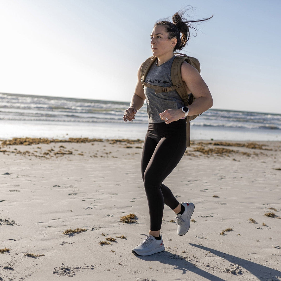 Jogging on a sandy beach with a backpack, they rely on the Rough Runner - Light Grey + Red from GORUCK, whose road-to-trail outsole easily adapts to the changing terrain. The ocean serves as a tranquil backdrop while the Gradient Density EVA midsole supports every stride.