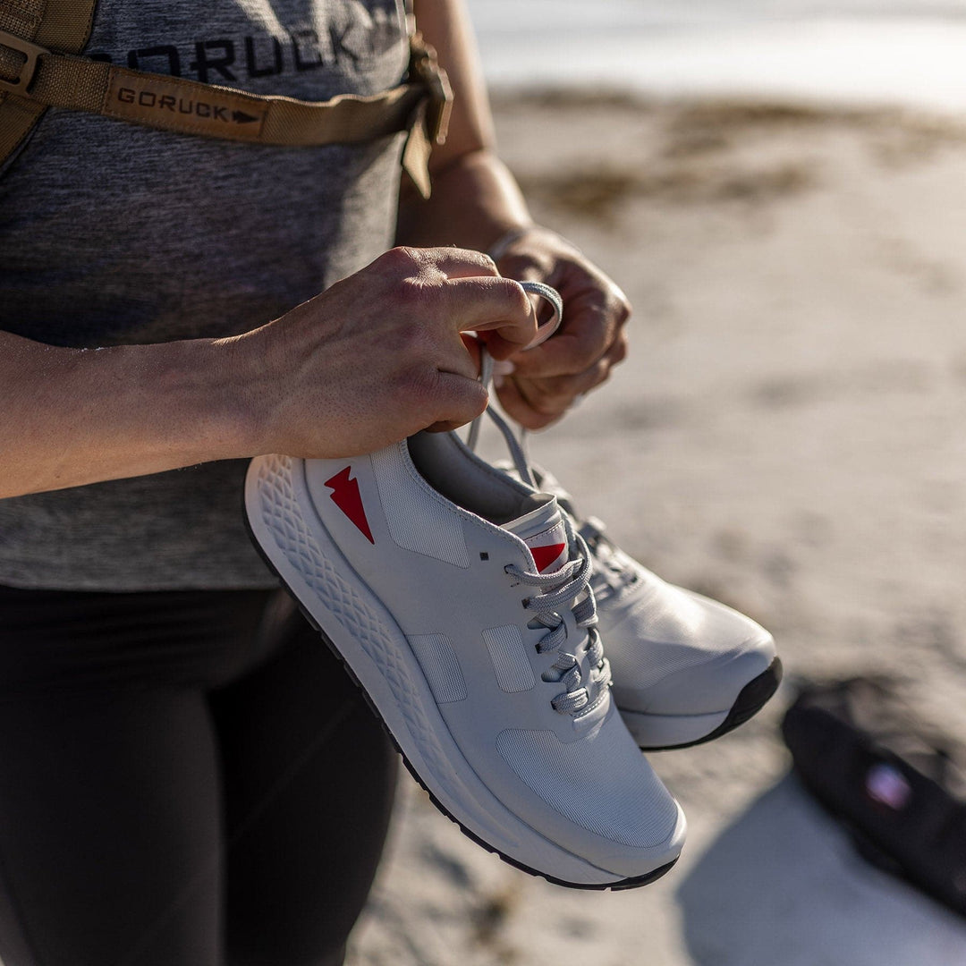 A person outdoors holds the Rough Runner shoes by GORUCK in a light grey color with red accents, highlighted by a Gradient Density EVA midsole designed for comfort.
