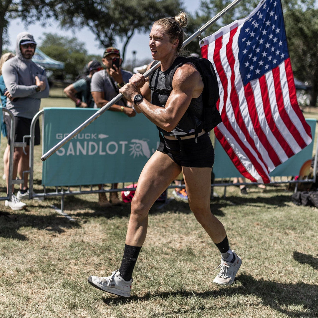 An athlete, proudly carrying a USA flag while running in a sports event on a grassy field, showcases their GORUCK Rough Runner - Light Grey + Red road-to-trail outsole running shoes.