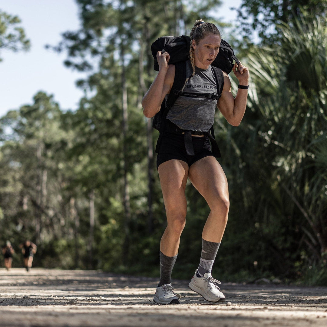 A person, wearing GORUCK's Rough Runner shoes in Light Grey + Red with a road-to-trail outsole, is carrying a heavy backpack while walking on a dirt path in a forested area.