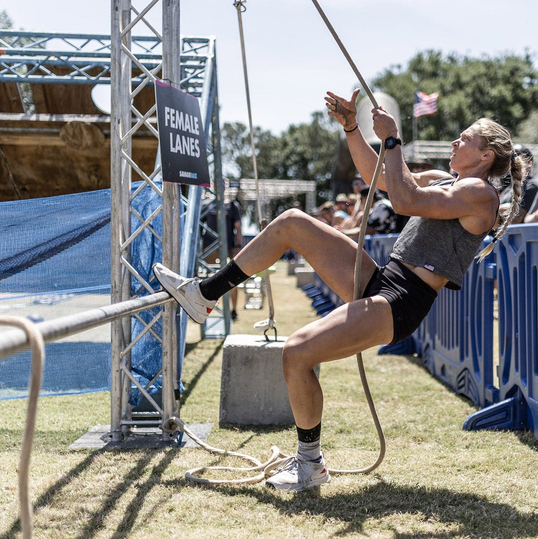 At an outdoor fitness event, a woman ascends a rope beside the "Female Lanes" sign, demonstrating her agility. She's sporting GORUCK's Rough Runner shoes in Light Grey + Red, designed with a road-to-trail outsole that makes them ideal for various terrains and confirms their adaptability in every challenge she encounters.