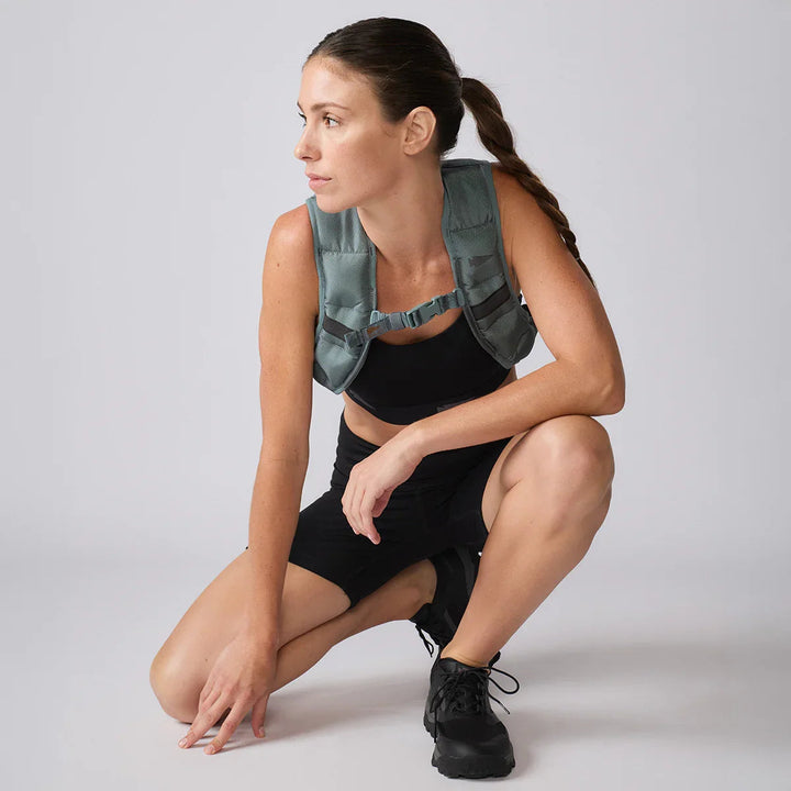 Woman in athletic wear and GORUCK weighted ruck vest squats on light background