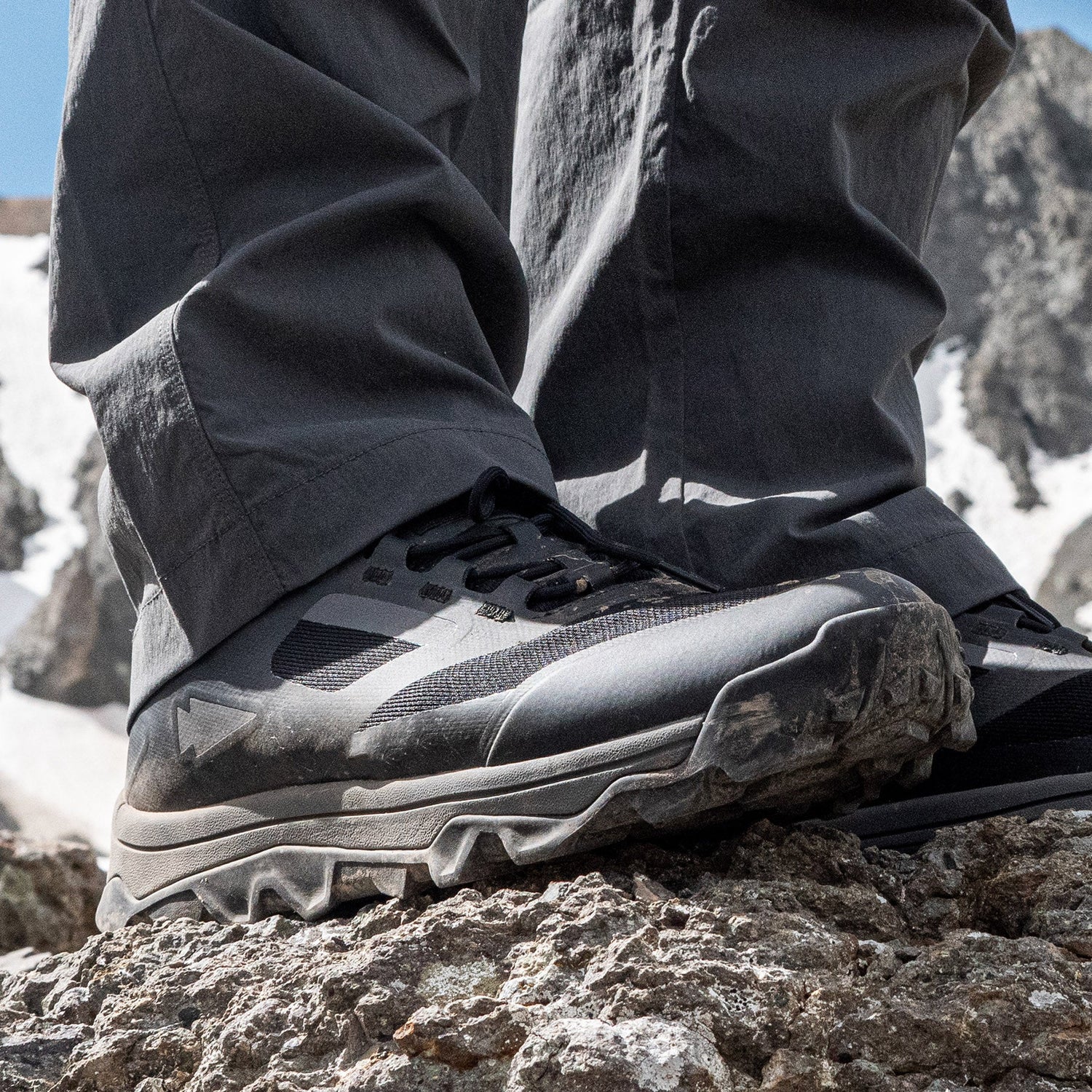Close-up of hiker’s muddy hiking shoes on rocky terrain with mountains and snow in the background.