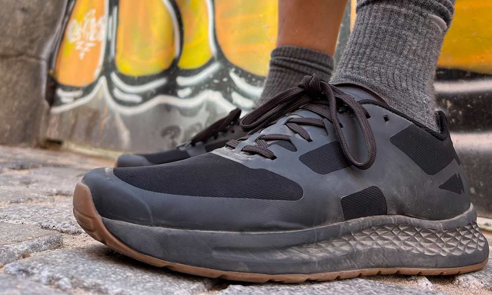 Person wearing black sneakers and gray socks standing on cobblestone pavement with graffiti in the background.