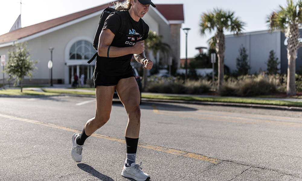 Person wearing athletic gear runs outdoors on a sunny street with a building and palm trees in the background.