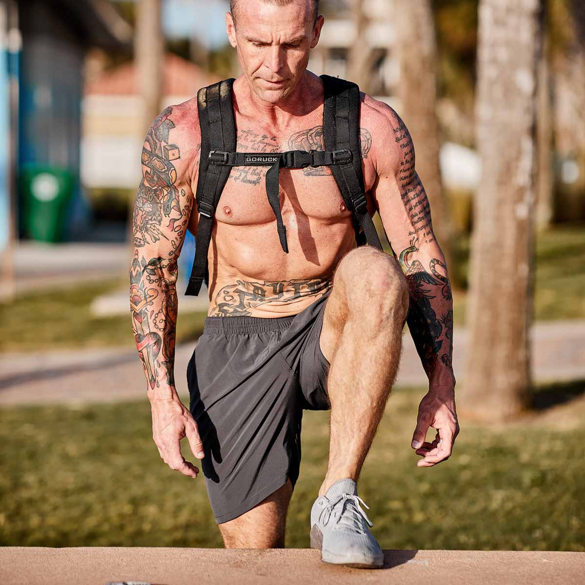 Tattooed man in workout gear and backpack does a step-up exercise outdoors in a sunny park.