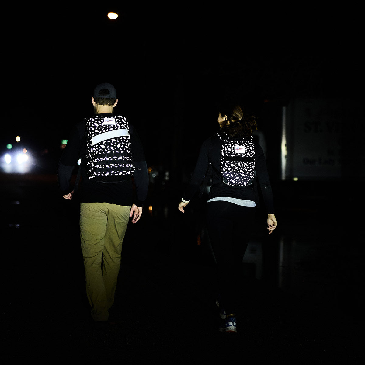 Two individuals wearing GORUCK Ruck Plate Carrier 3.0 packs are walking at night, illuminated by headlights against a dark background. These carriers provide ergonomic lumbar support and include a padded top handle, ensuring a comfortable and efficient midnight trek.