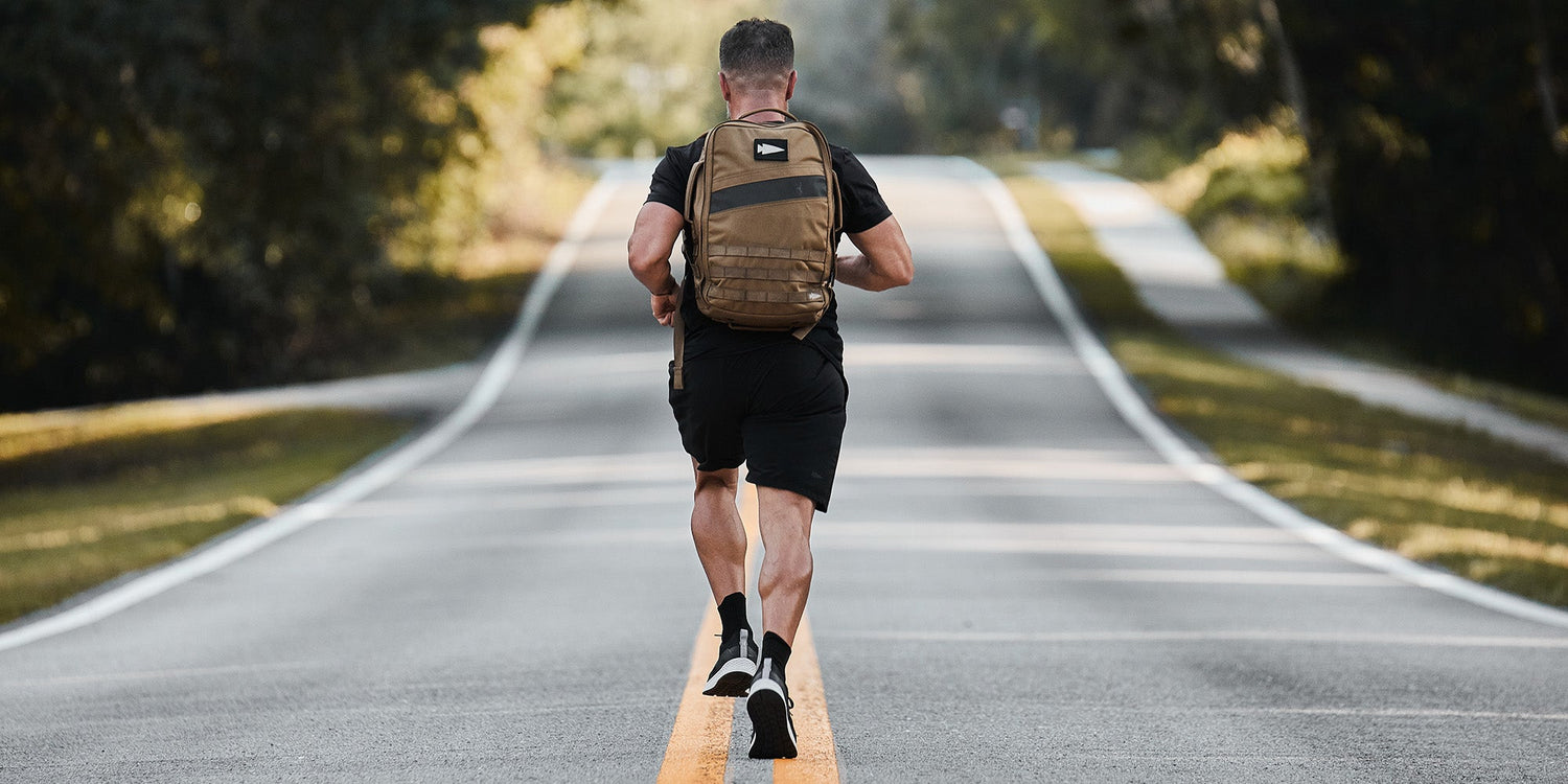 Man jogging down a rural road wearing a backpack, surrounded by trees and sunlight.