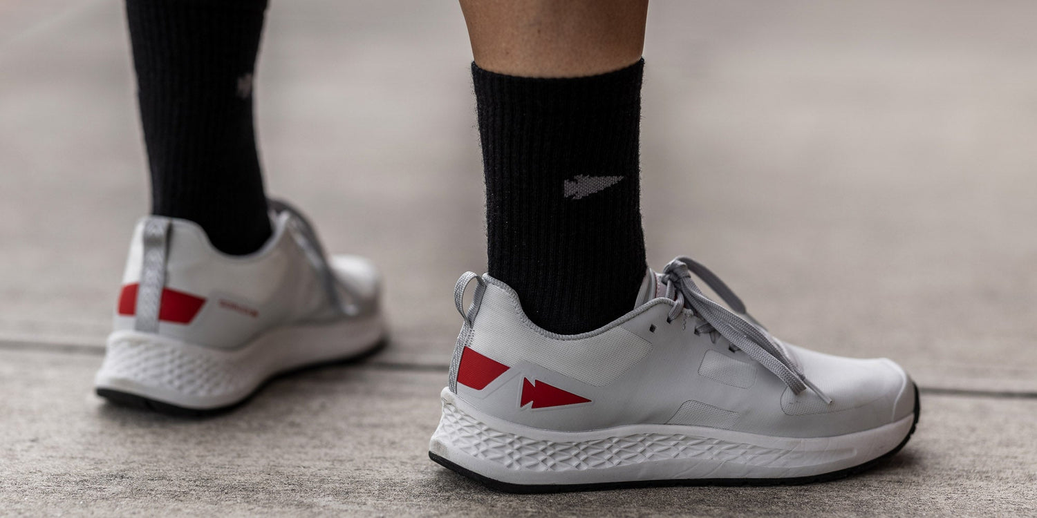 White athletic shoes with red accents and black socks, worn by a person standing on a concrete surface.