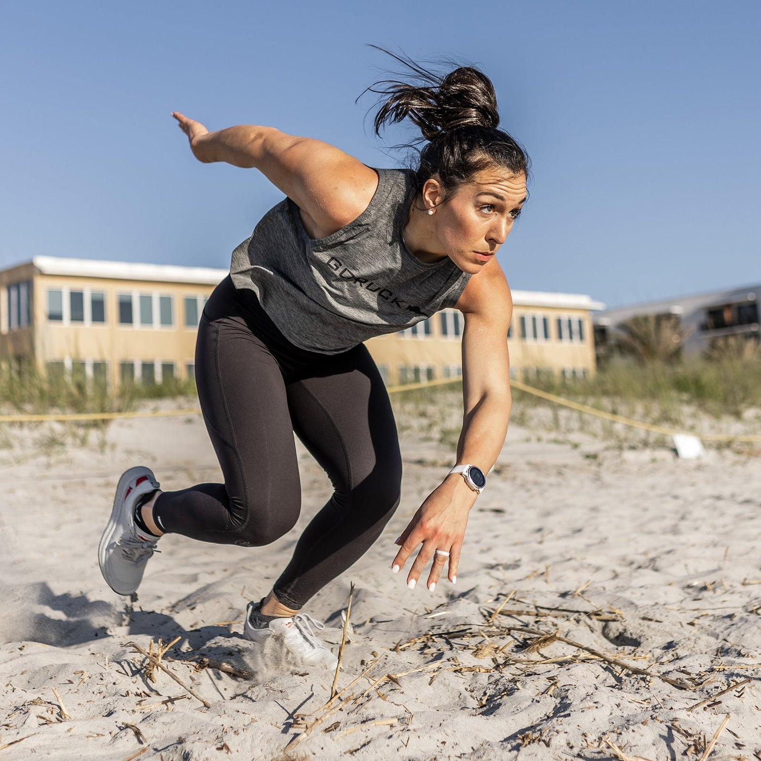 Woman in athletic wear sprinting on a sandy beach, looking focused, with buildings in the background.