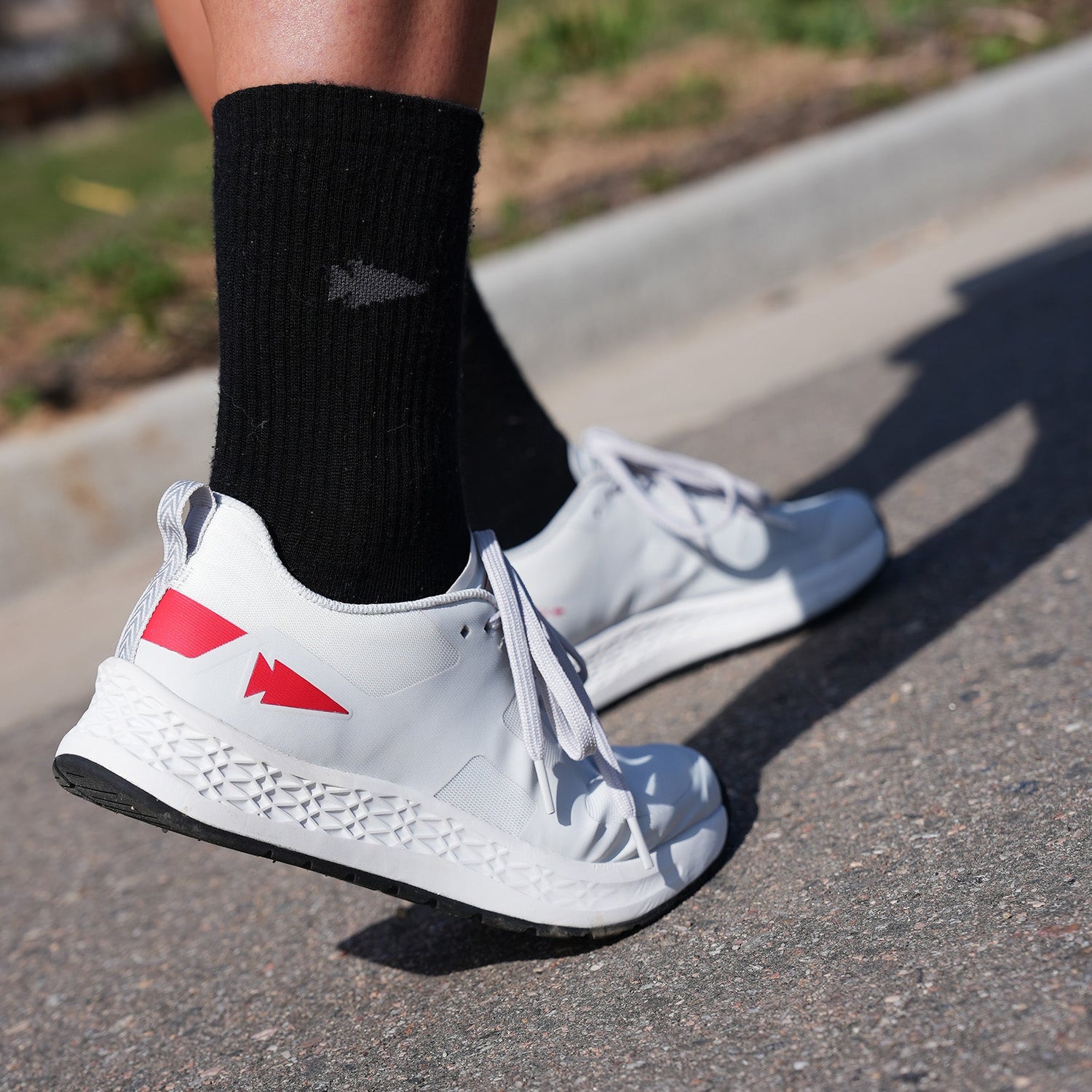 Person wearing white running shoes with red accents and black socks, standing on a paved surface outdoors.