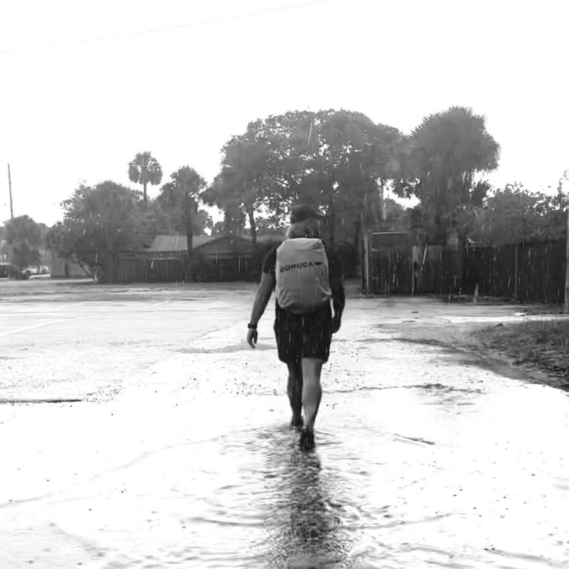 Person walking alone in the rain on a flooded street, wearing a backpack, with trees in the background.