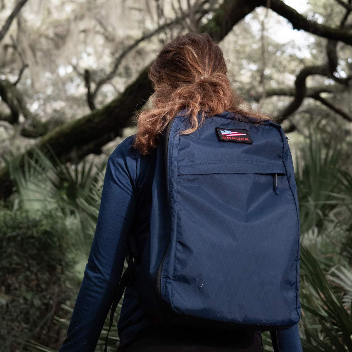 Person with a blue backpack stands in a forest, facing away toward trees and greenery.