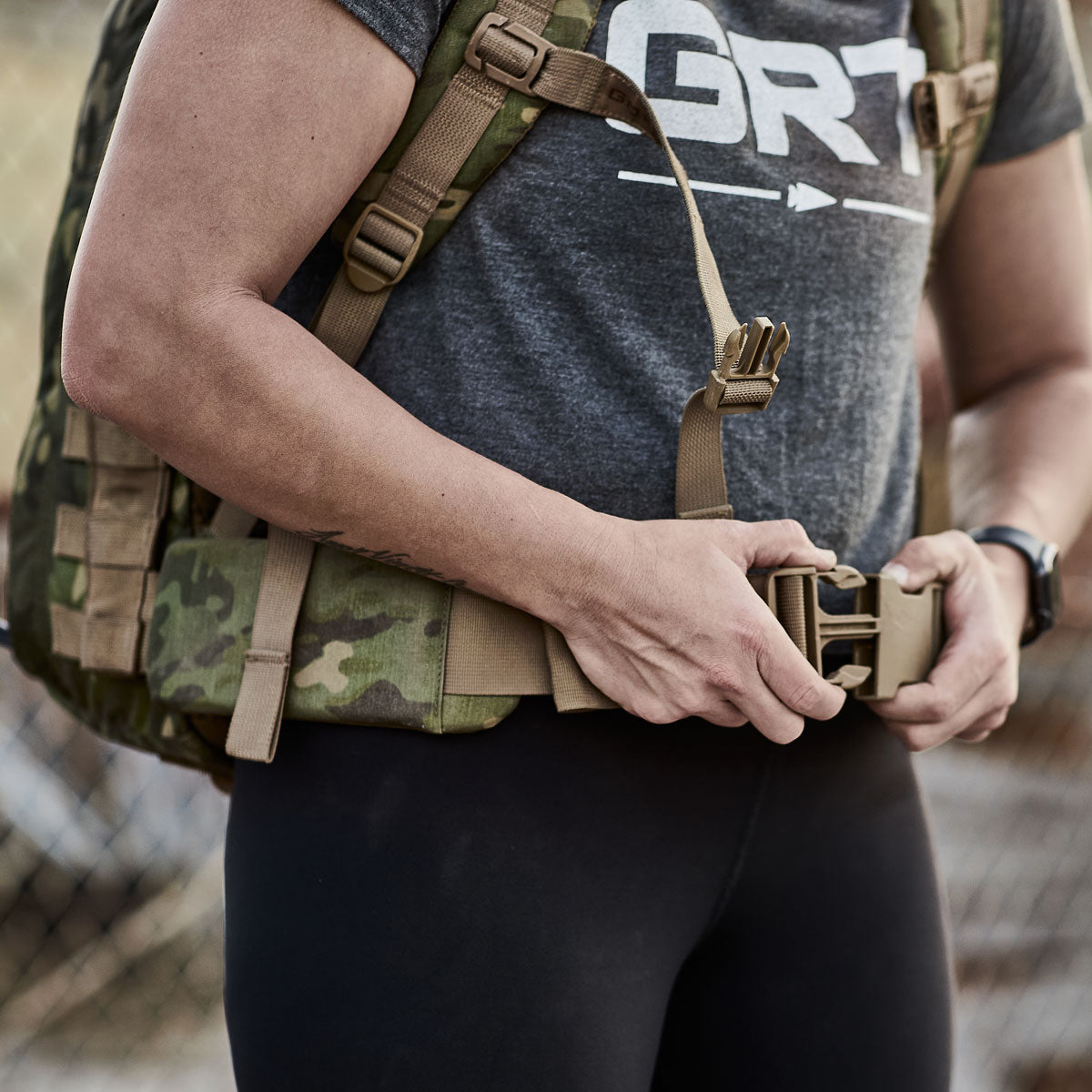 Person wearing a grey t-shirt buckling a camouflage backpack waist strap outdoors.