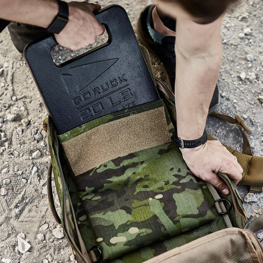 A person places a 20 lb GORUCK weight plate into a camouflage backpack on gravel.