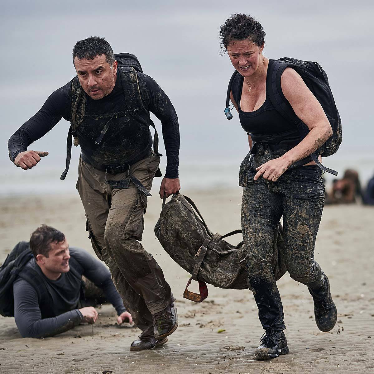 Two muddy people in athletic gear run on a beach, carrying a heavy sandbag, with another person behind them.