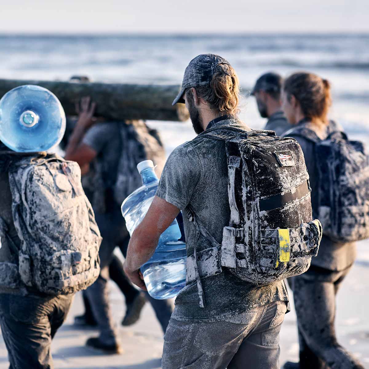 A group of people in muddy gear carry water jugs and a log while walking along a sandy beach.