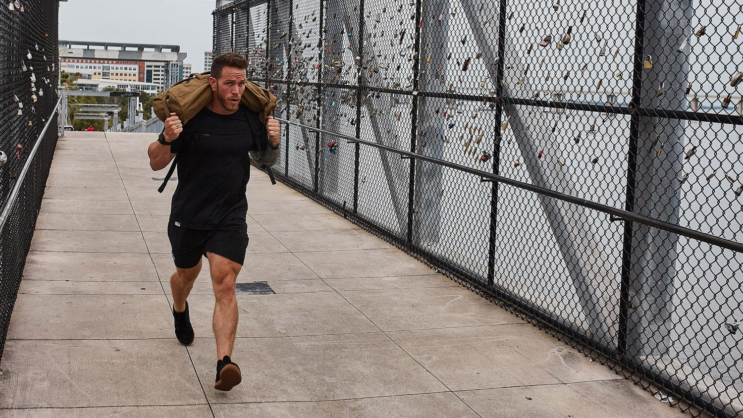 Man running on a bridge, carrying a heavy sandbag on his shoulders, with city buildings in the background.