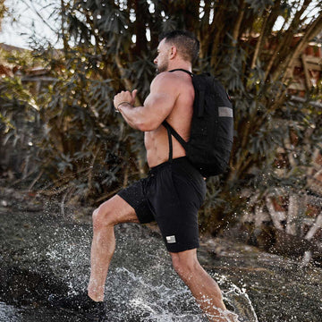 Shirtless man with a black backpack runs through shallow water, splashing, with trees in the background.
