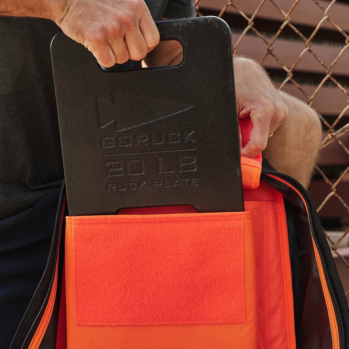 A person places a 20-pound black ruck plate, featuring a carved GORUCK logo and weight markings, into an orange Rucker 4.0 backpack. Ideal for rucking enthusiasts, the scene is set against a quiet chain-link fence in the background.