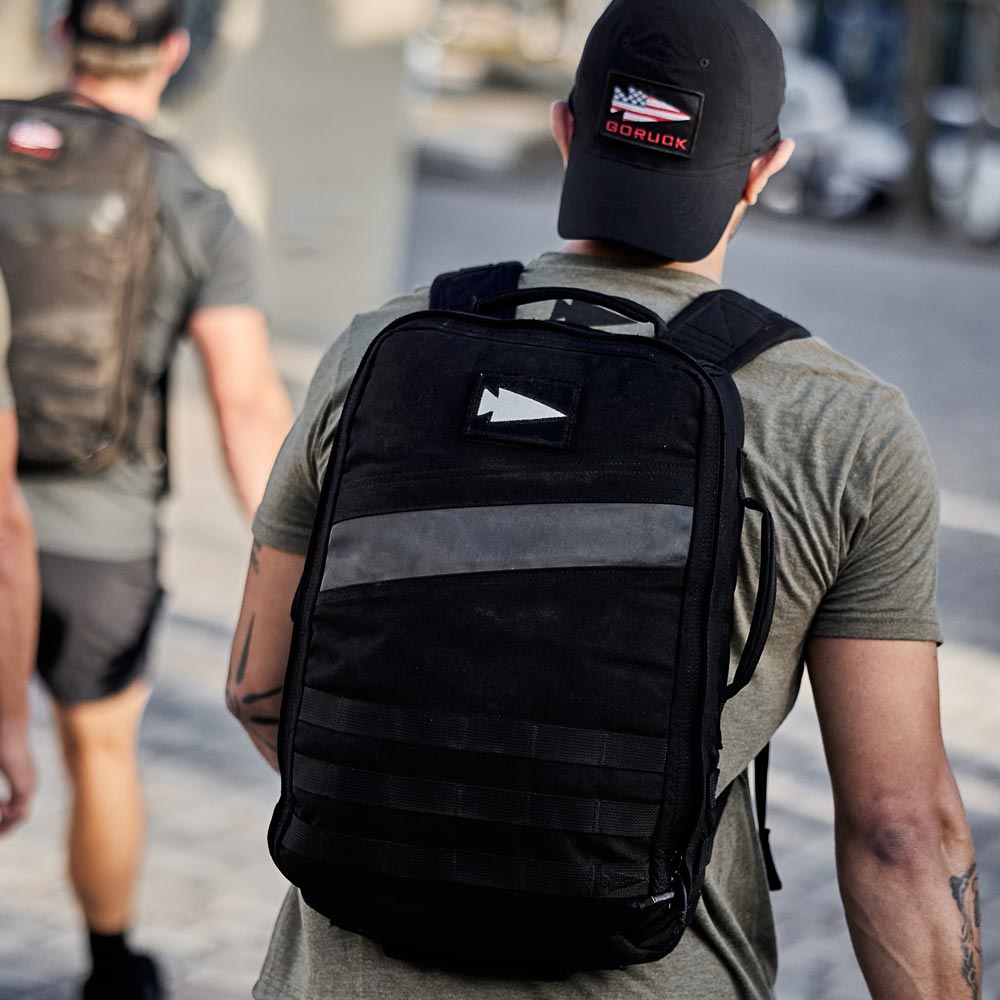 Person wearing a black backpack and cap walking outdoors, another person with a backpack in front of them.