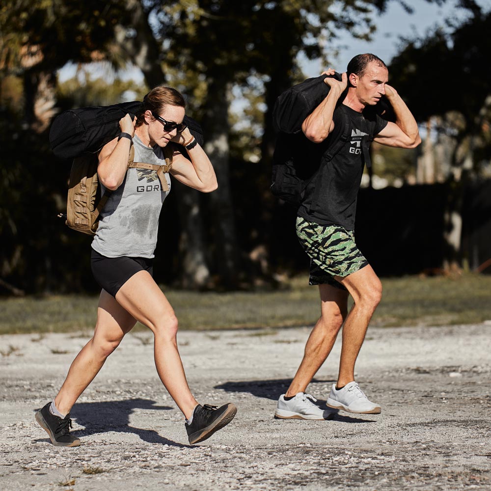Two people exercise outdoors, carrying heavy bags on their shoulders while walking on a dirt path.