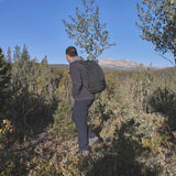 A person wearing a black jacket and GORUCK Men's Simple Pants - Midweight ToughDry® with a backpack stands on a rocky hill, surrounded by trees and foliage. In the background, there is a clear blue sky and a distant mountain peak.