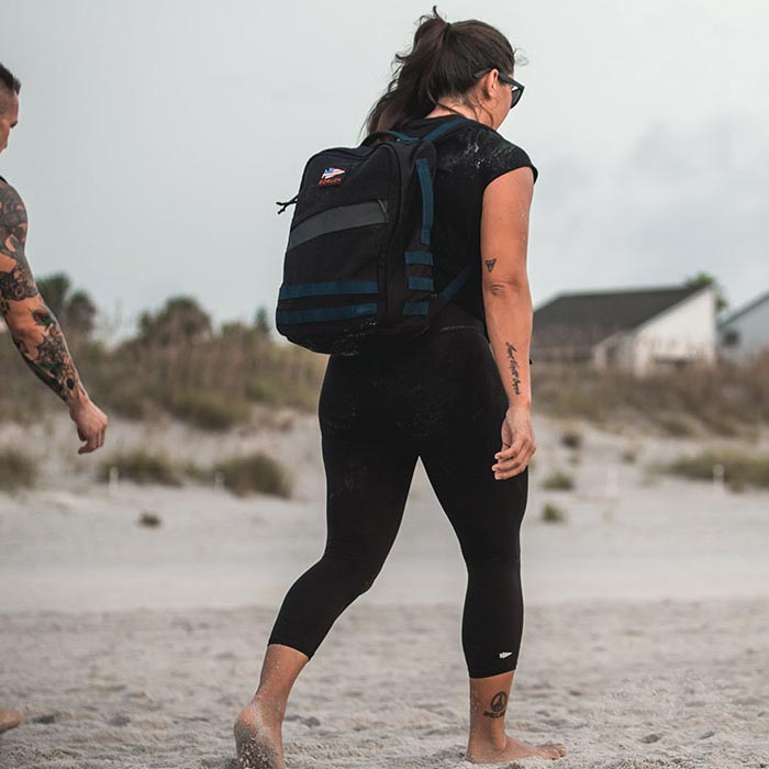 Woman in black leggings and backpack walks barefoot on a sandy beach, with another person behind her.