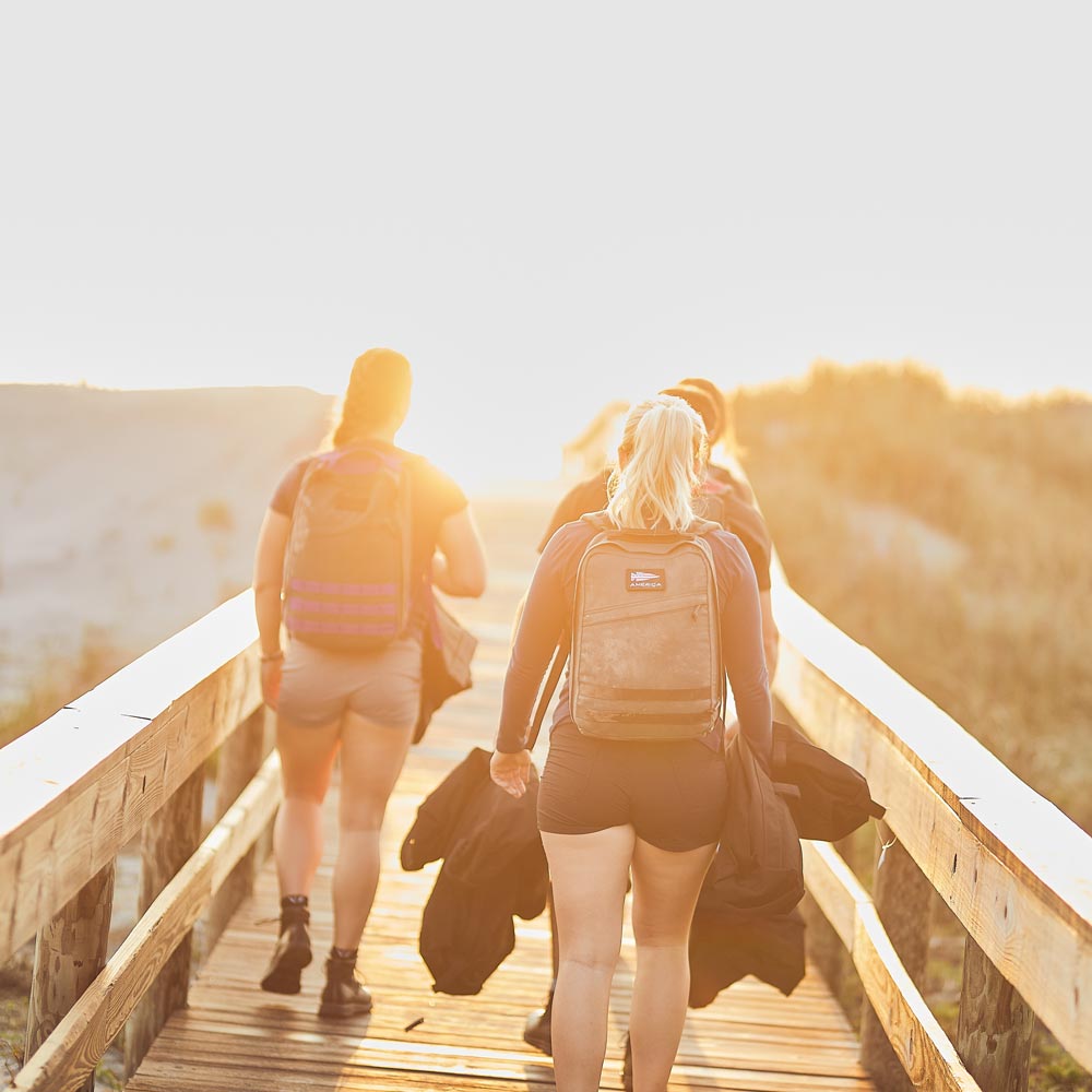 Three people with backpacks walk on a wooden path toward the sunlight, surrounded by nature.