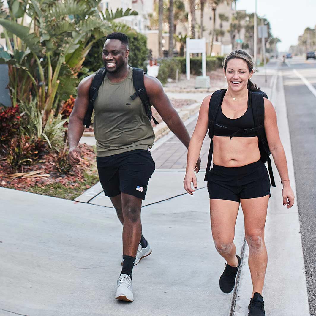 Two people wearing athletic clothes and backpacks smile while walking outside on a sunny day.