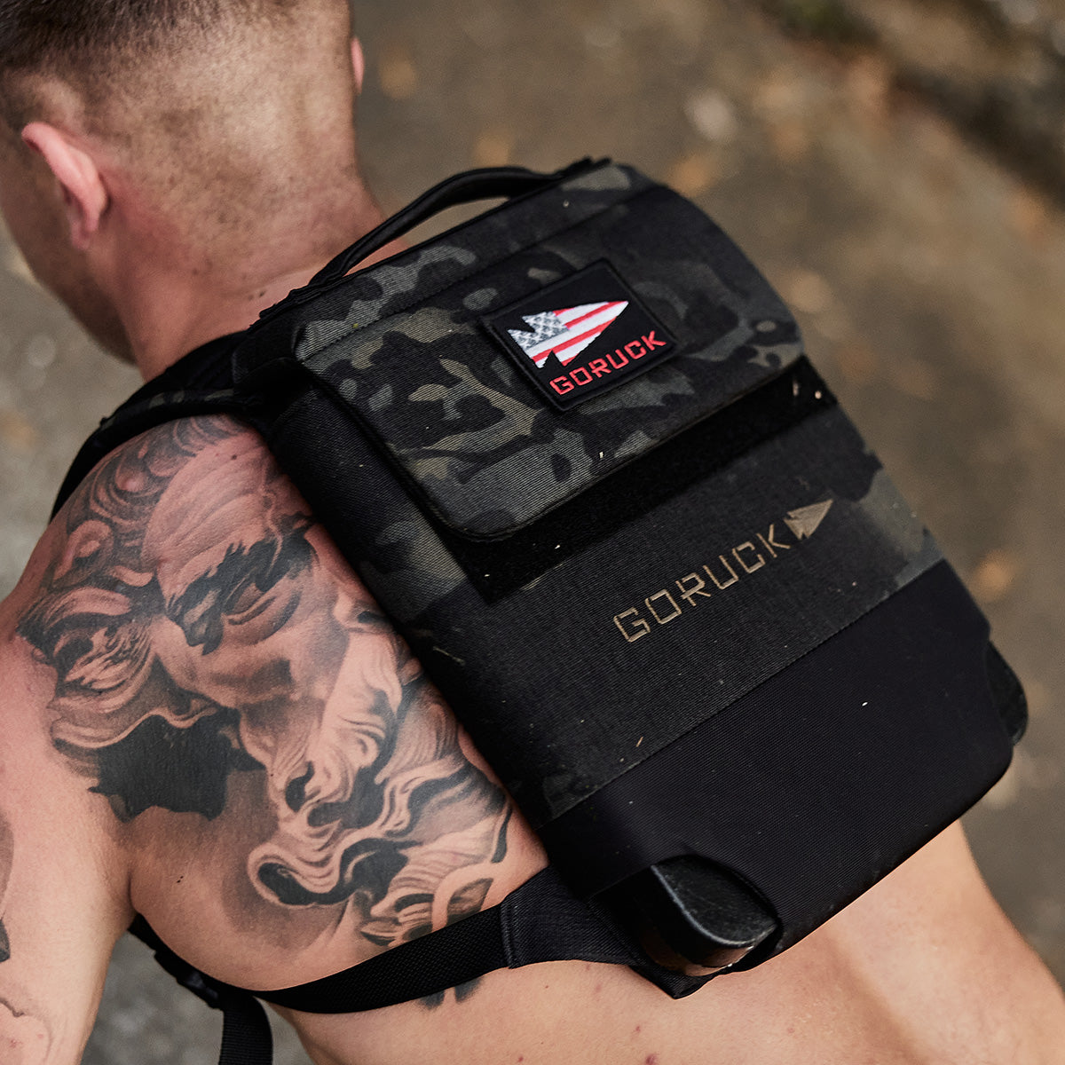 A tattooed man wearing a black GORUCK backpack with a US flag patch on his shoulder.