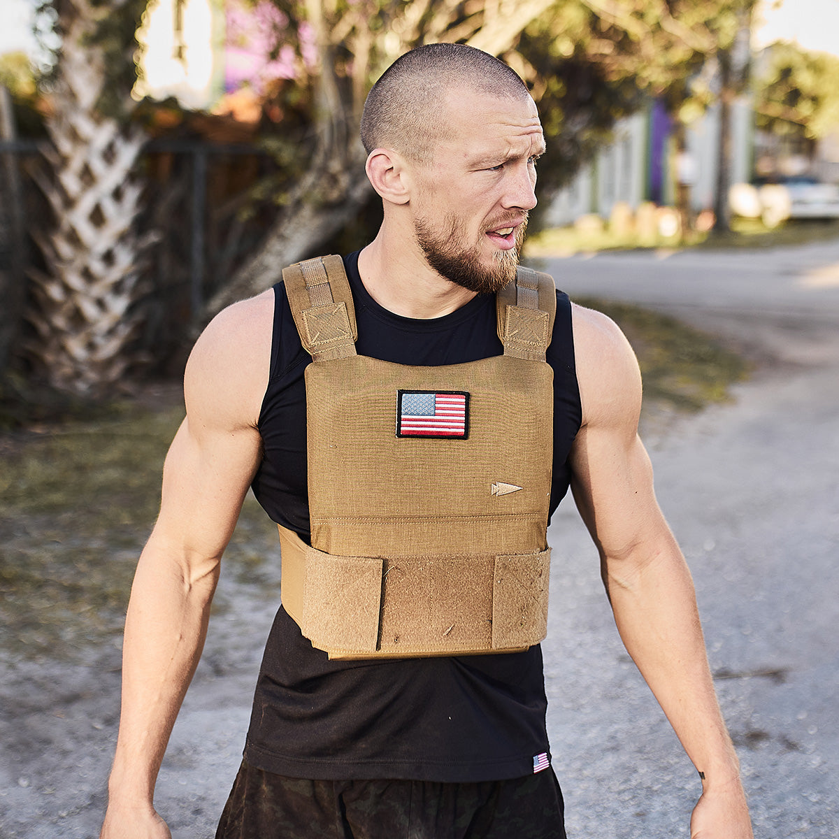 A man wearing a tan tactical vest with a U.S. flag patch stands outdoors, looking to his left.