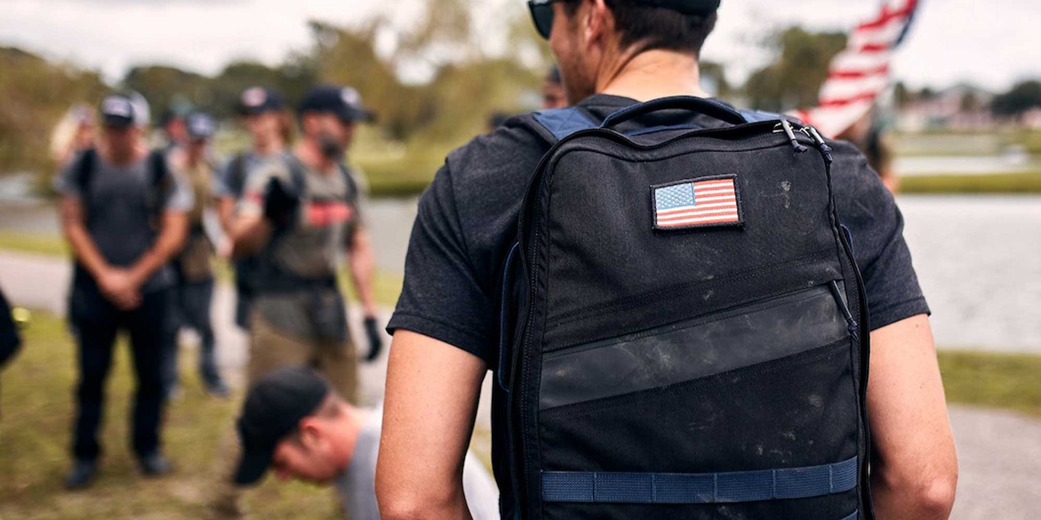 Person wearing a backpack with a U.S. flag patch, standing among a group outdoors near water.