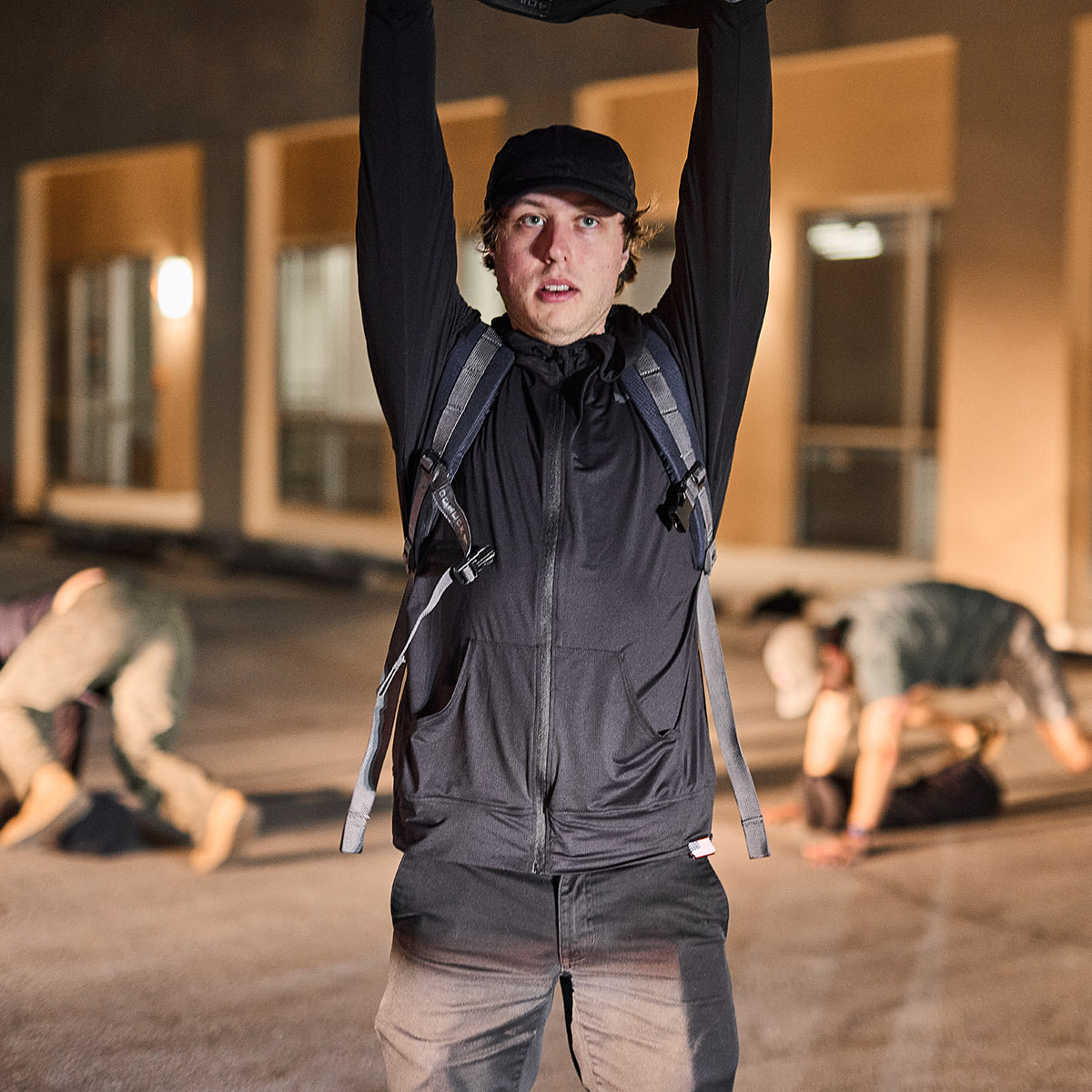 Man in black hoodie lifts object overhead outside at night, with two people exercising in the background.