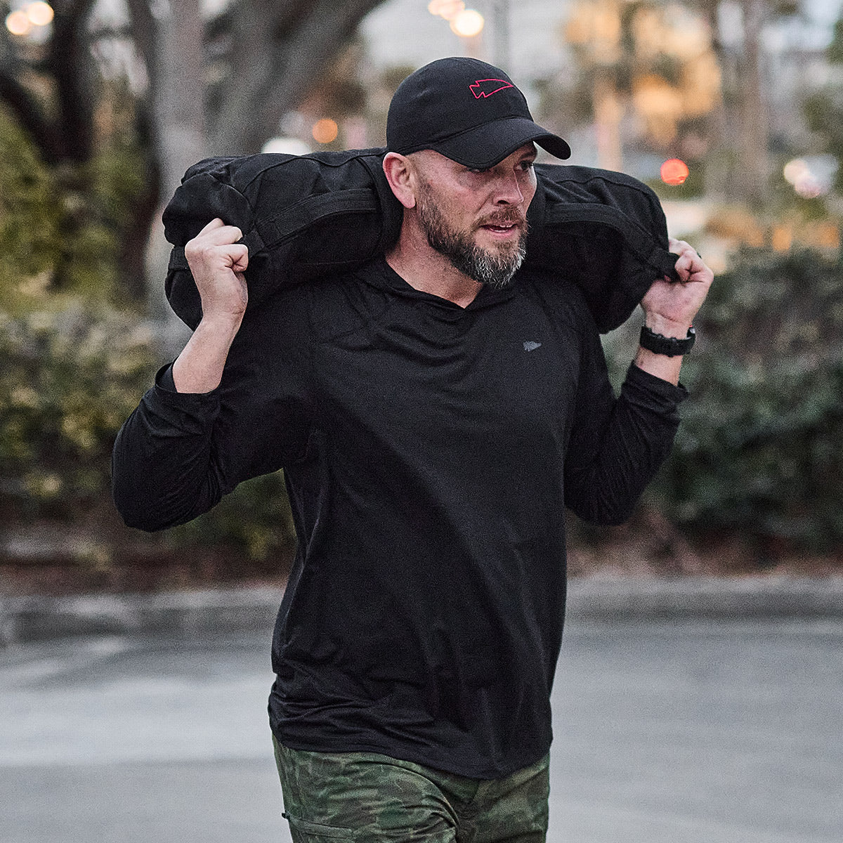 Man in black shirt and cap carrying a heavy sandbag on his shoulders outdoors.