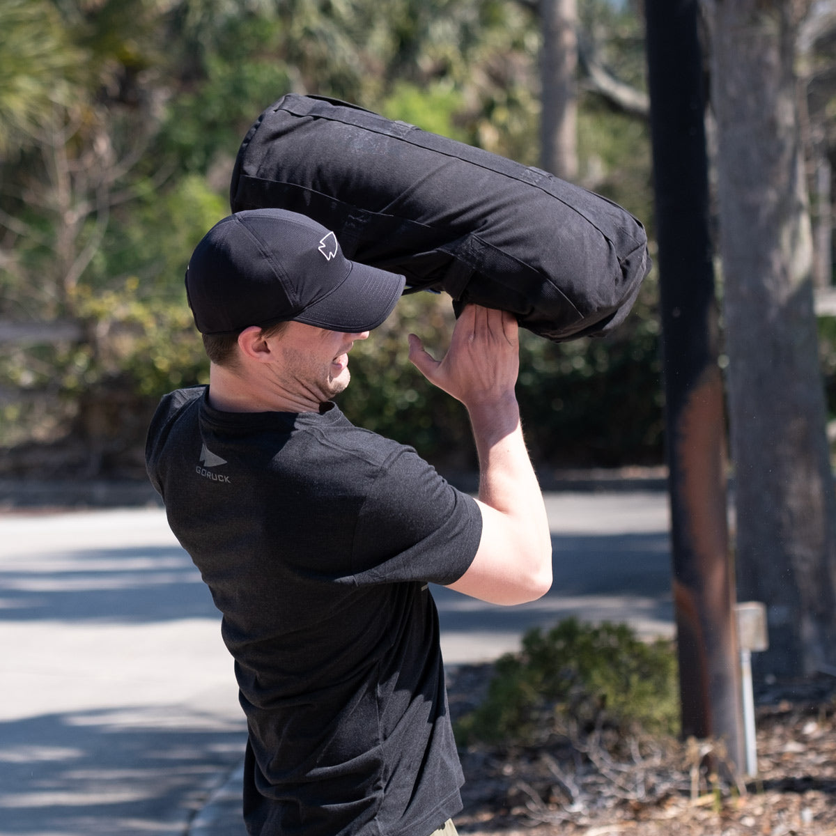 A man in a black shirt and hat lifts a large black sandbag outdoors on a sunny day.