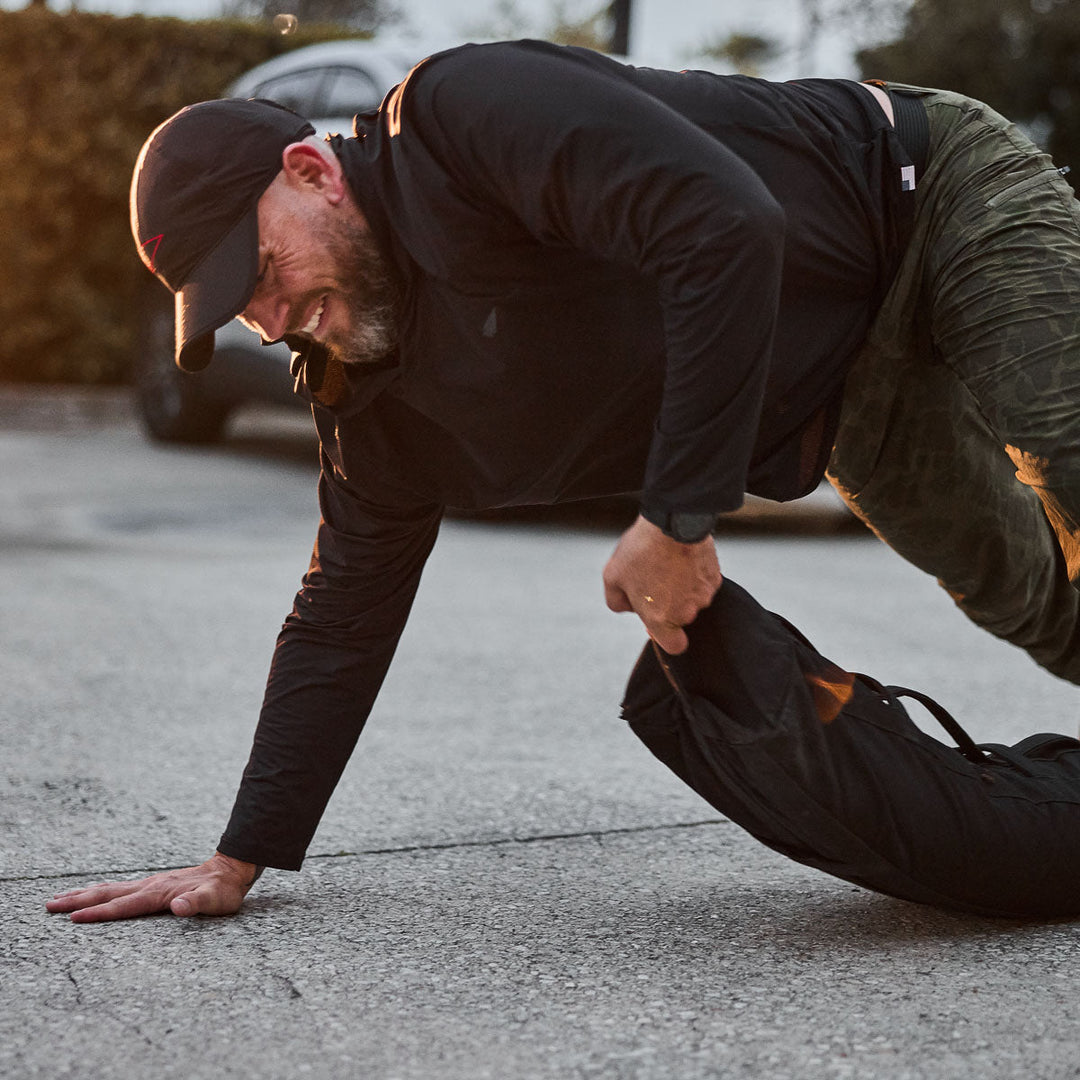 Wearing the GORUCK Performance Running Hat - ToughMesh, a man does push-ups outdoors on concrete, gripping a heavy sandbag.