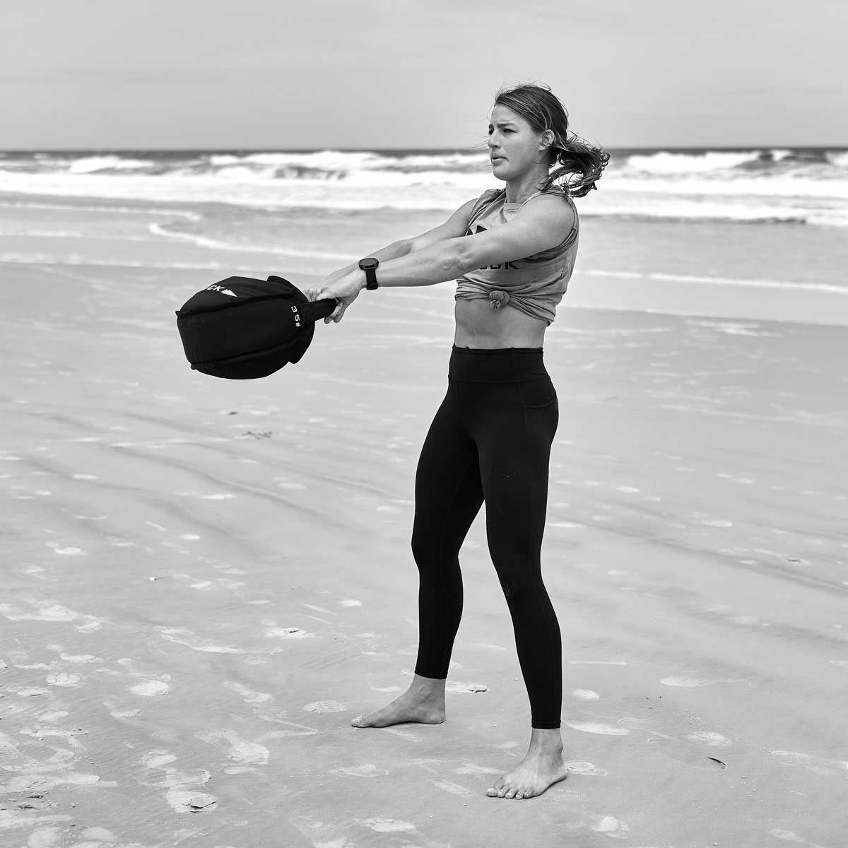 A woman exercises with a sandbag on a sandy beach by the ocean, wearing athletic clothing.
