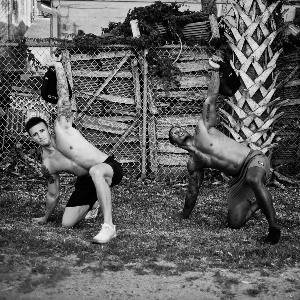 Two shirtless men outdoors doing one-arm kettlebell exercises on grass by a chain-link fence.